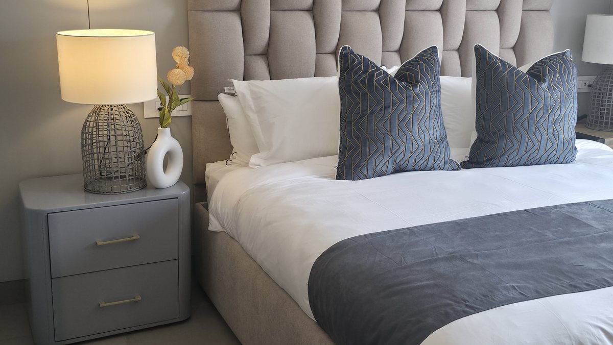 Bed with a grey tufted headboard, blue patterned cushions and a lit bedside lamp.