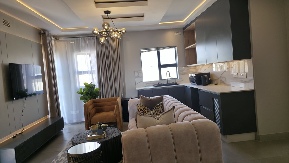 Living area with a curved grey sofa, armchair, wall-mounted television and open kitchenette.