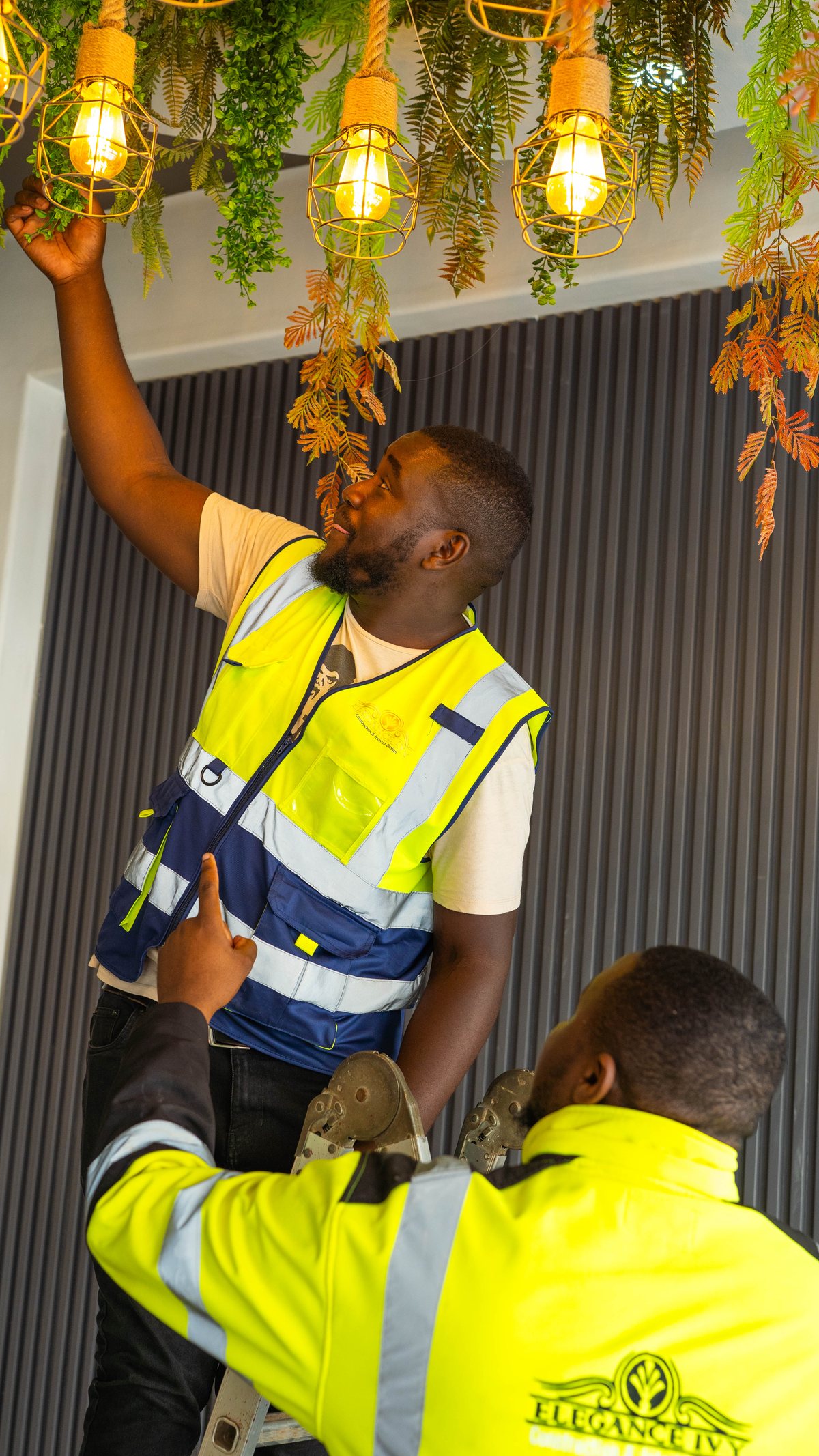 Two workers in reflective vests working beneath pendant lights and hanging foliage.