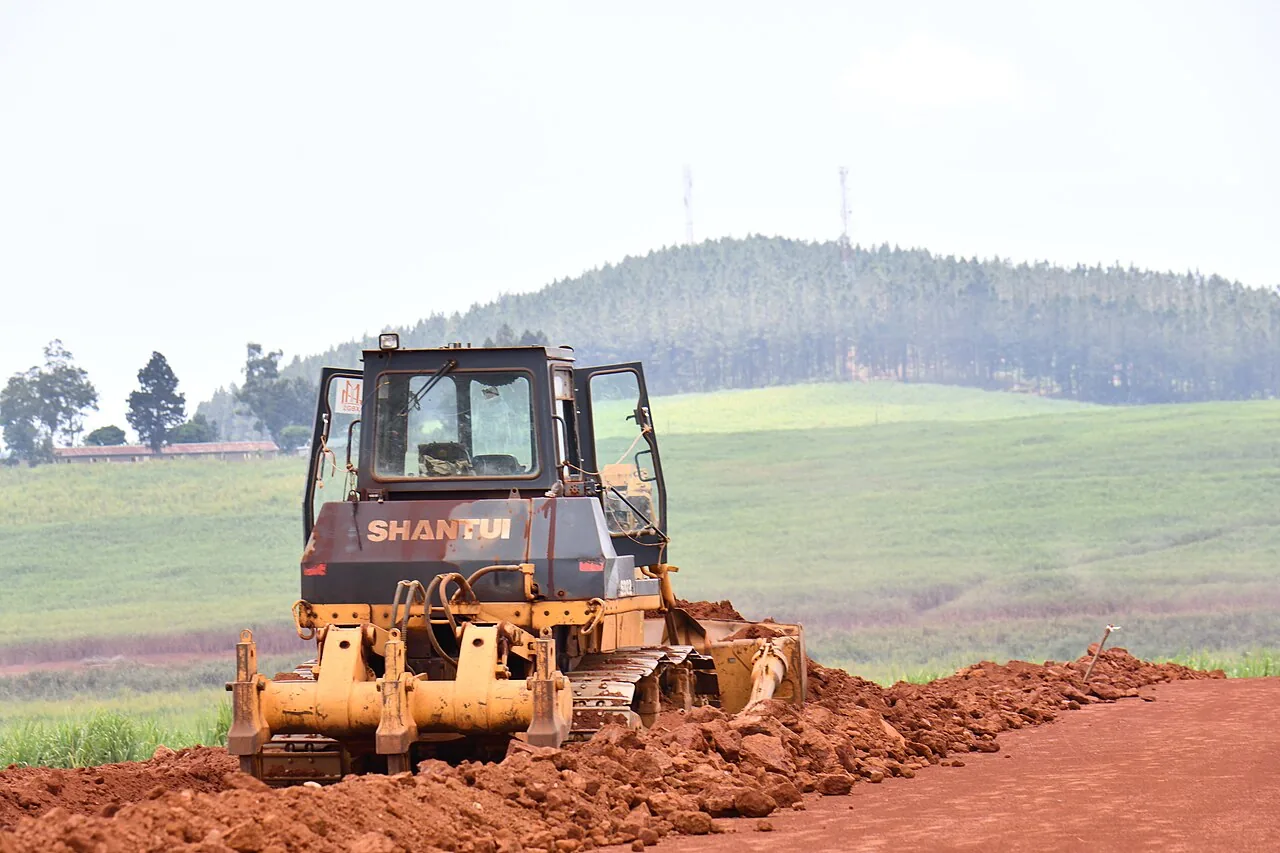 Tracked earthmoving machine grading a red-soil access route beside green fields.