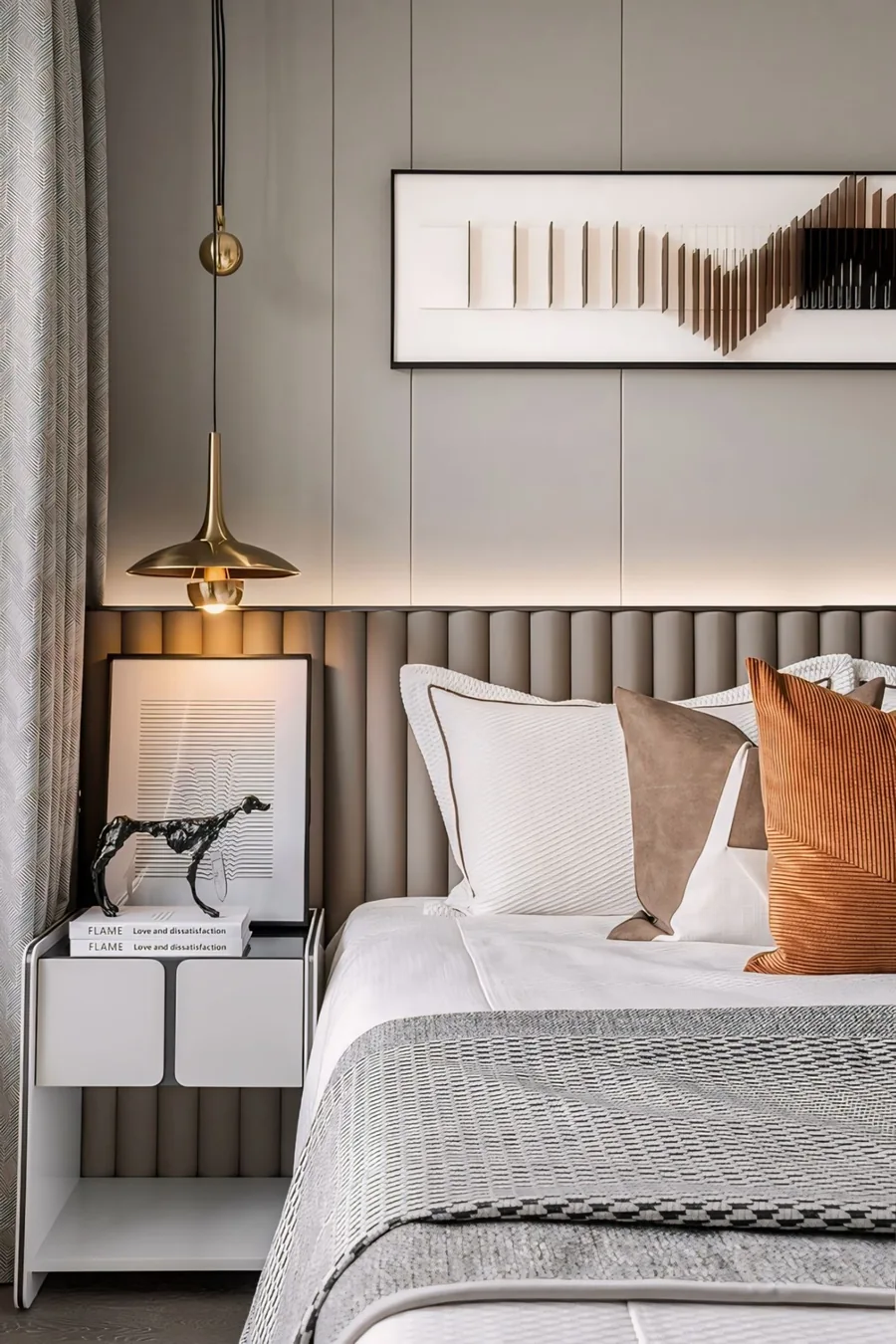 Bedroom detail with a padded headboard, hanging brass lights, framed artwork and layered cushions.